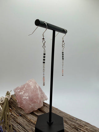 A pair of long beaded dangle earrings displayed on a black jewelry stand, with a pink crystal and dried stems in the background against a light gray backdrop.