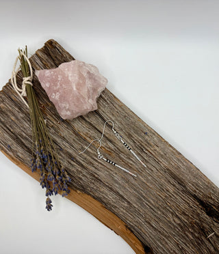 A pair of silver beaded drop earrings displayed on a rustic piece of driftwood, alongside a pink crystal and a small bundle of dried lavender stems, against a plain white background.