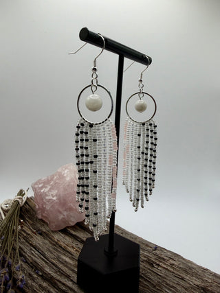 A pair of long fringe earrings displayed on a black stand. Each earring has a silver-tone hoop with a round rainbow moonstone bead, followed by multiple strands of clear and black seed beads hanging in a fringe style.