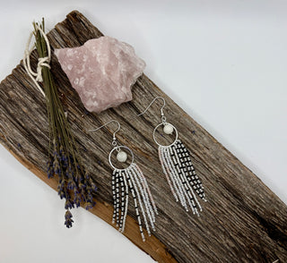 A pair of silver fringe earrings with circular hoops, rainbow moonstone beads, and long strands of black-and-white seed beads, displayed on a rustic wood slice beside a pink crystal and dried lavender stems.