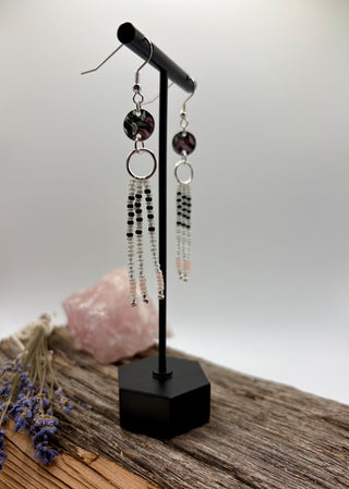A pair of long beaded dangle earrings displayed on a black jewelry stand. The earrings feature a dark purple round bead, a silver ring, and cascading strands of black and clear beads.