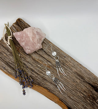 Silver dangle earrings with circular charms and beaded fringe, displayed on a rustic wood slice beside a pink crystal and dried lavender stems against a white background.
