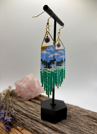 A pair of handmade beaded dangle earrings displayed on a black stand. The earrings feature a whale tail coming out of the ocean, suspended from gold-toned metal frames with small purple beads.