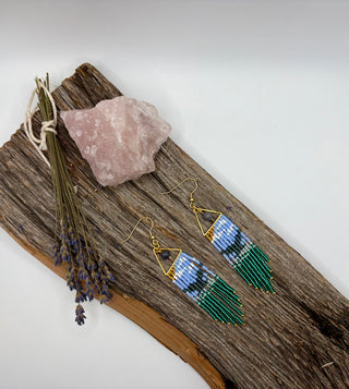 A pair of handmade beaded dangle earrings displayed on a rustic piece of wood, alongside a pink crystal and dried lavender sprigs, against a clean white background. The beads form the pattern of a whale tail in the ocean.