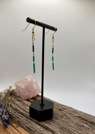 A pair of long beaded dangle earrings displayed on a black T-stand. The earrings feature gold fishhook wires, small black and white bead sections, and green beaded drops.