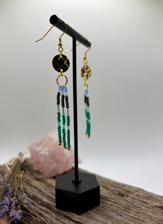 A pair of bohemian-style beaded dangle earrings displayed on a black jewelry stand. The earrings feature a round black and white marbled disc, a gold ring, a textured gold disc, and three strands of beads in light blue, black, and green.