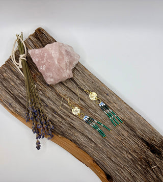 A pair of handmade bohemian dangle earrings displayed on a rustic piece of driftwood alongside a pink crystal and dried lavender. The earrings feature gold-plated hooks and circular charms, with one dangling tassel made of white beads and another made of green beads with white and black accents.