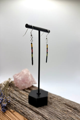 A pair of long beaded drop earrings displayed on a black T-stand, set on a rustic wooden surface with a pink crystal and dried lavender in the background.