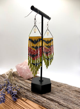 A pair of long beaded fringe earrings displayed on a black stand. Long seed bead fringe earrings featuring bats on a yellow, carnelian, and yellow background with moonstone accents.