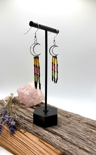 A pair of long beaded dangle earrings with silver crescent moon hoops, displayed on a black stand. The earrings feature vertical strands of black, yellow, red, and green beads.