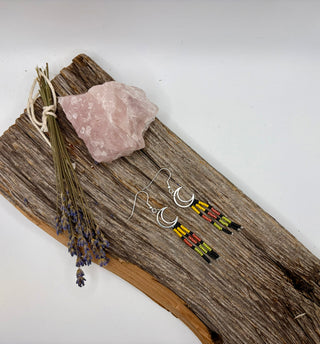 A pair of silver crescent moon dangle earrings with colorful beaded fringe, displayed on a rustic piece of wood alongside a pink crystal and dried lavender stems against a plain white background.