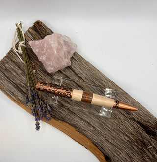 A decorative copper and wood pen displayed on a rustic piece of driftwood with a pink crystal and dried lavender sprigs, photographed against a plain white background.