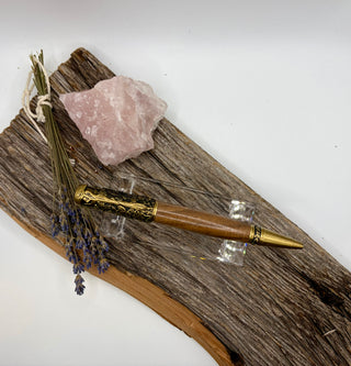 A wooden and brass pen with ornate gold-toned detailing, displayed on a piece of weathered driftwood alongside a pink crystal, dried lavender, and a small bundle of white string.