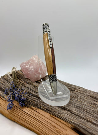 A wooden pen with silver metal accents displayed on a clear acrylic stand, set on a piece of driftwood with a pink crystal and dried lavender sprigs against a light gray background.