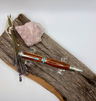 A decorative wooden pen with antique pewter metal accents and turquoise details, displayed on a piece of driftwood with a pink crystal and dried lavender sprigs against a white background.