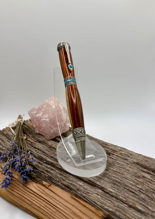 A wooden pen with antique pewter accents and turquoise blue inlay details, displayed on a clear acrylic stand on a piece of weathered wood next to a pink crystal and dried lavender.