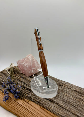 A Sapele wood pen with chrome metal accents stands upright on a clear acrylic display stand. The setup is styled on a piece of weathered wood with a pink crystal and dried lavender sprigs against a neutral gray background.