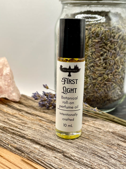 Roll-on perfume oil bottle labeled 'First Light' on a wooden surface with lavender and a crystal.