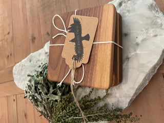 A set of solid hardwood Black Walnut coasters with a tag showing the product name and a raven logo, placed on a natural stone surface with green foliage in the background.