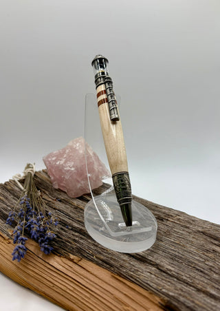 A Maple wood pen withe bloodwood inlays with pewter-toned metal accents displayed on a clear acrylic stand on a piece of weathered wood with a pink crystal and dried lavender.