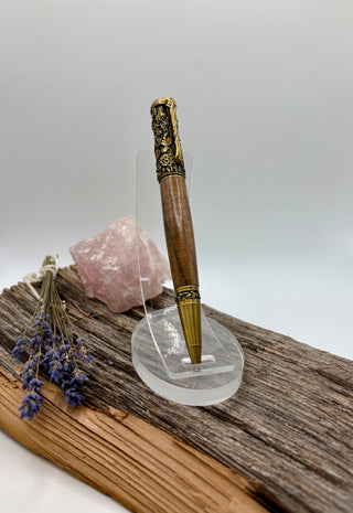 A black walnut pen with ornate antique brass accents stands on a clear acrylic display stand. The pen is styled on a piece of weathered wood next to a pink crystal cluster and dried lavender sprigs against a neutral gray background.