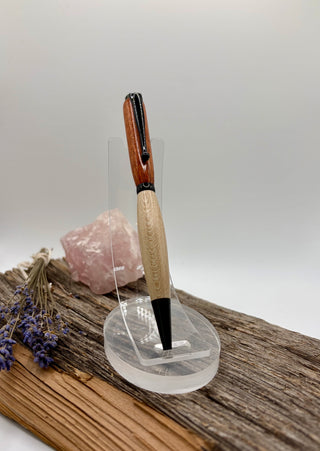 Got to catch them all!  A wooden pen with a bloodwood top and a light beige middle section, displayed on a clear acrylic stand. The pen is set on a piece of weathered wood next to a pink crystal and dried lavender sprigs against a neutral gray background.