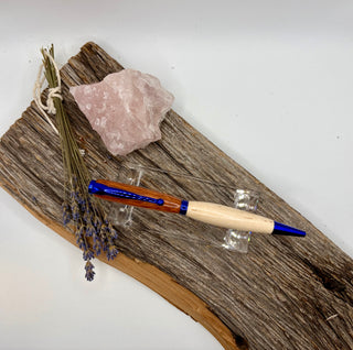 A wooden and metal pen with blue accents rests on a rustic piece of driftwood alongside a pink crystal and dried lavender sprigs, photographed against a plain white background.