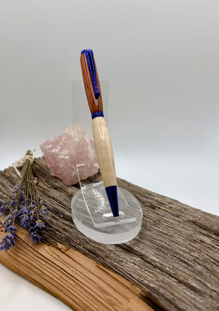 A wooden pen with blue hardware with a maple bottom and bloodwood top stands on a clear acrylic display stand, placed on a rustic piece of wood with a pink crystal and dried lavender sprigs against a light gray background.