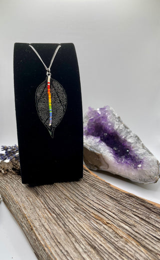 Leaf-shaped pendant with rainbow design on a black card, placed on a wooden surface with a purple crystal in the background.