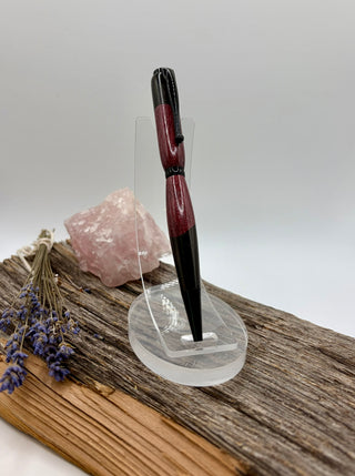 A two-tone pen with black and deep purple sections, displayed on a clear acrylic stand on a piece of weathered wood with a pink crystal and dried lavender.