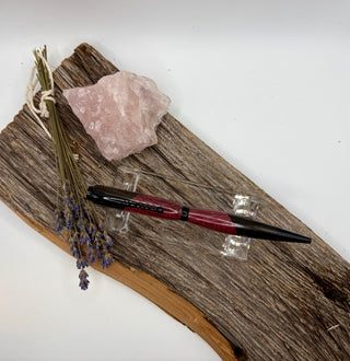 A deep purple and black pen displayed on a rustic piece of driftwood with a pink crystal and dried lavender stems, photographed against a plain white background.