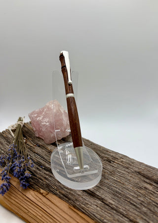 A wooden Sapele pen with satin chrome accents displayed on a clear acrylic stand, set on a rustic wood slice with a pink crystal and dried lavender in the background.