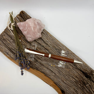 A wooden pen with silver metal accents displayed on a clear stand, resting on a piece of weathered driftwood alongside a pink crystal and dried lavender sprigs, against a plain white background.