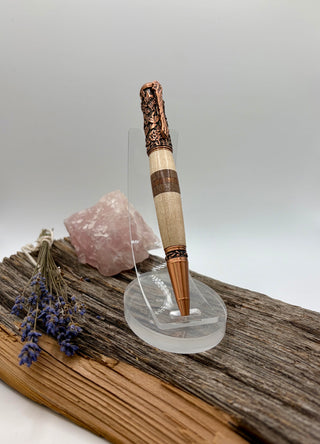An ornate botanical ambrosia maple, black walnut and sapele pen with a carved antique copper-finished top, a light wood handle, and dark brown bands, displayed on a clear acrylic stand. The stand is placed on a piece of driftwood with a pink crystal and dried lavender in the background against a neutral gray studio backdrop.