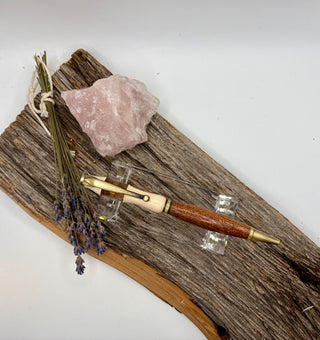 A phenomenal wooden pen with a brown wood-grain barrel and cream-colored accents, displayed on a piece of driftwood alongside a pink crystal, dried lavender, and a string bundle against a plain white background.