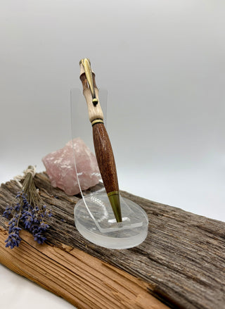 The most beautiful carved wooden pen money can buy with metallic gold accents, displayed on a clear acrylic stand on a rustic wood surface with dried lavender and a pink crystal in the background.
