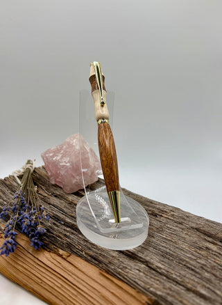 A wooden and gold pen with gold-tone metal accents, displayed on a clear acrylic stand on a rustic wood slice with a pink crystal and dried lavender.