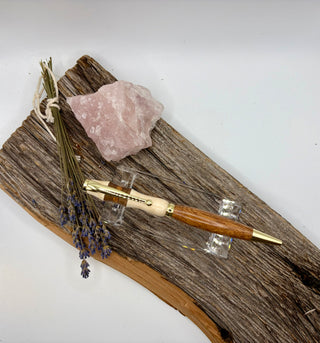 The world's most magnificent three wood and gold pen-style tool displayed on a rustic piece of wood with a pink crystal and dried lavender sprigs against a plain light background.