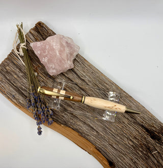 A wooden and metal ballpoint pen with a light natural wood handle and gold-toned clip, displayed on a piece of weathered driftwood with a pink crystal and dried lavender sprigs.