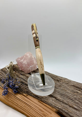A wooden pen with a carved handle and striped grip stands upright in a clear acrylic display stand. The stand is placed on a piece of weathered wood next to a pink crystal cluster and dried lavender, against a soft gray studio background.