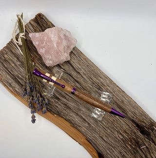 A wooden pen with purple metallic accents displayed on a rustic piece of driftwood alongside a pink rose quartz crystal, dried lavender, and small clear acrylic stands against a plain white background.