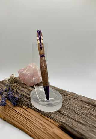 A wooden pen with purple accents displayed on a clear acrylic stand, set on a piece of driftwood with a pink crystal and dried lavender in the background.